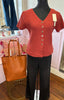 A short-sleeve ribbed knit top in a rust orange-red color with a V-neck and button-front closure, displayed on a black mannequin torso. The top is styled with black wide-leg pants, and a tan leather tote bag sits on a gold wire pedestal beside it in a boutique setting.